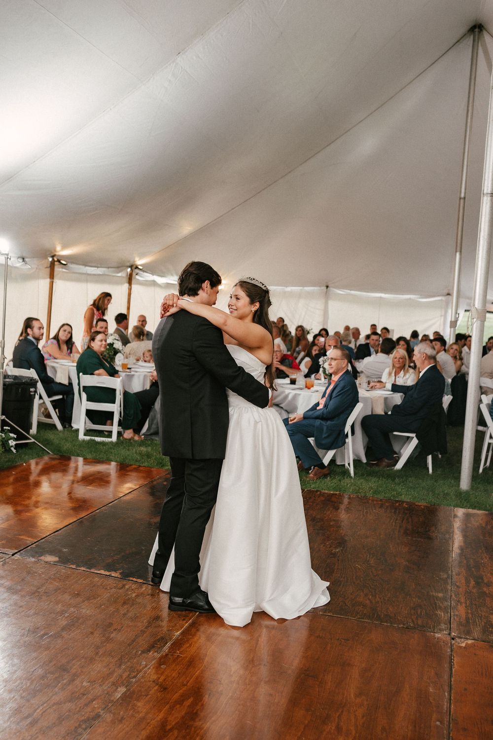 Bride and groom share their first dance on a wooden floor under a white tent with wedding guests seated at tables.
