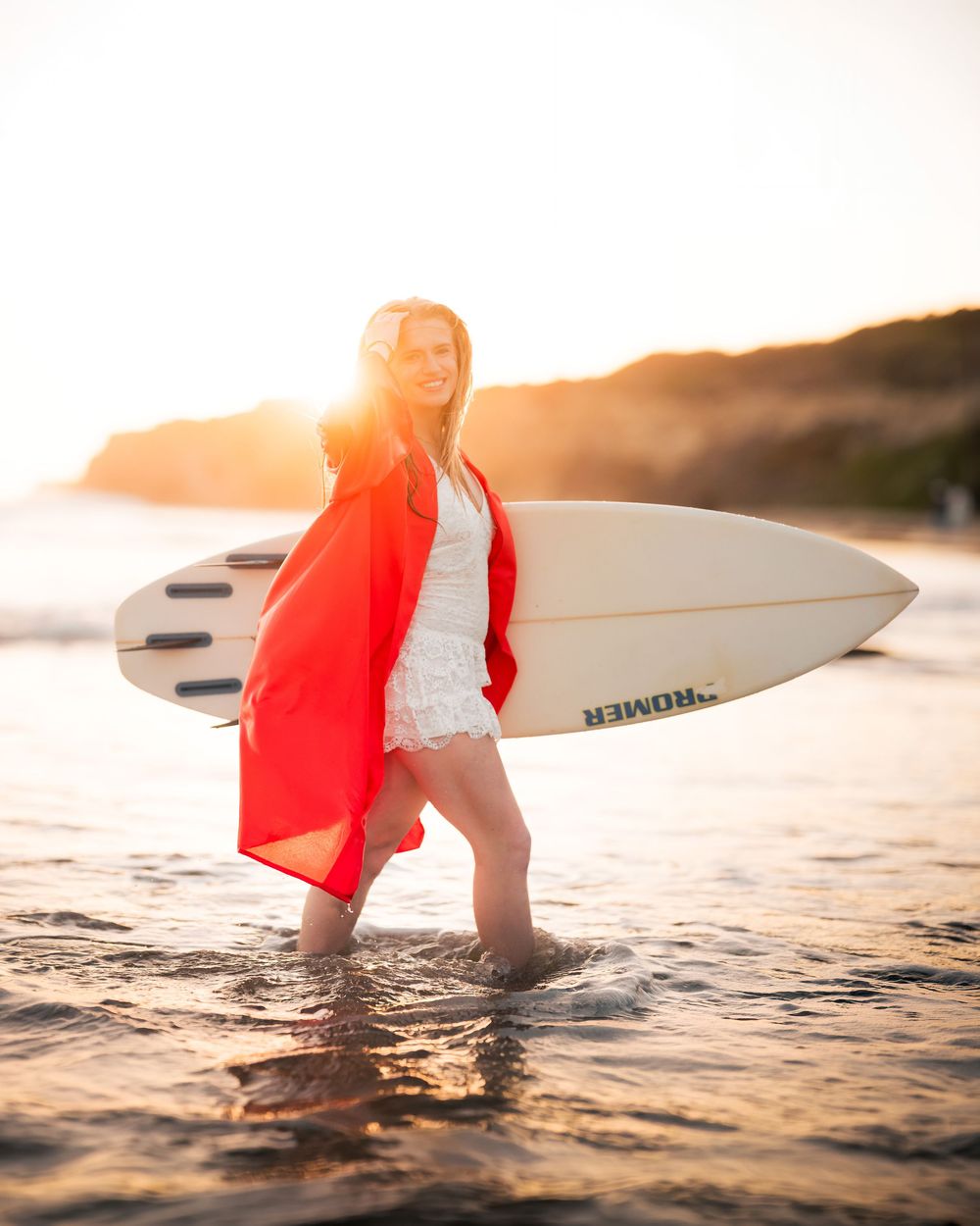 a female graduate in an orange cap and gown holding her surfboard next to the water in Crystal Cove, CA