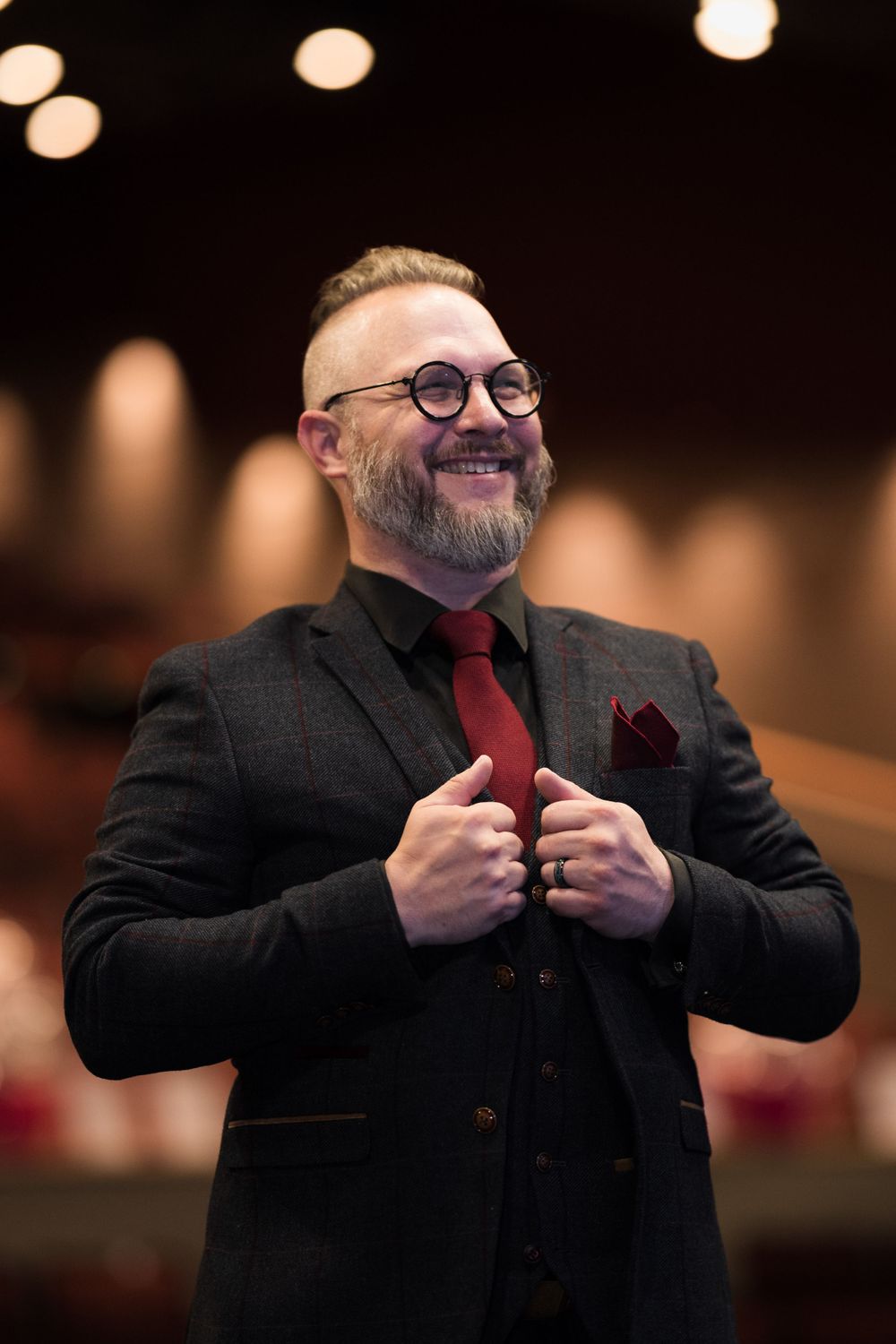 Bearded man in glasses wearing a gray and red suit, smiling in a theater with bokeh lights in the background.