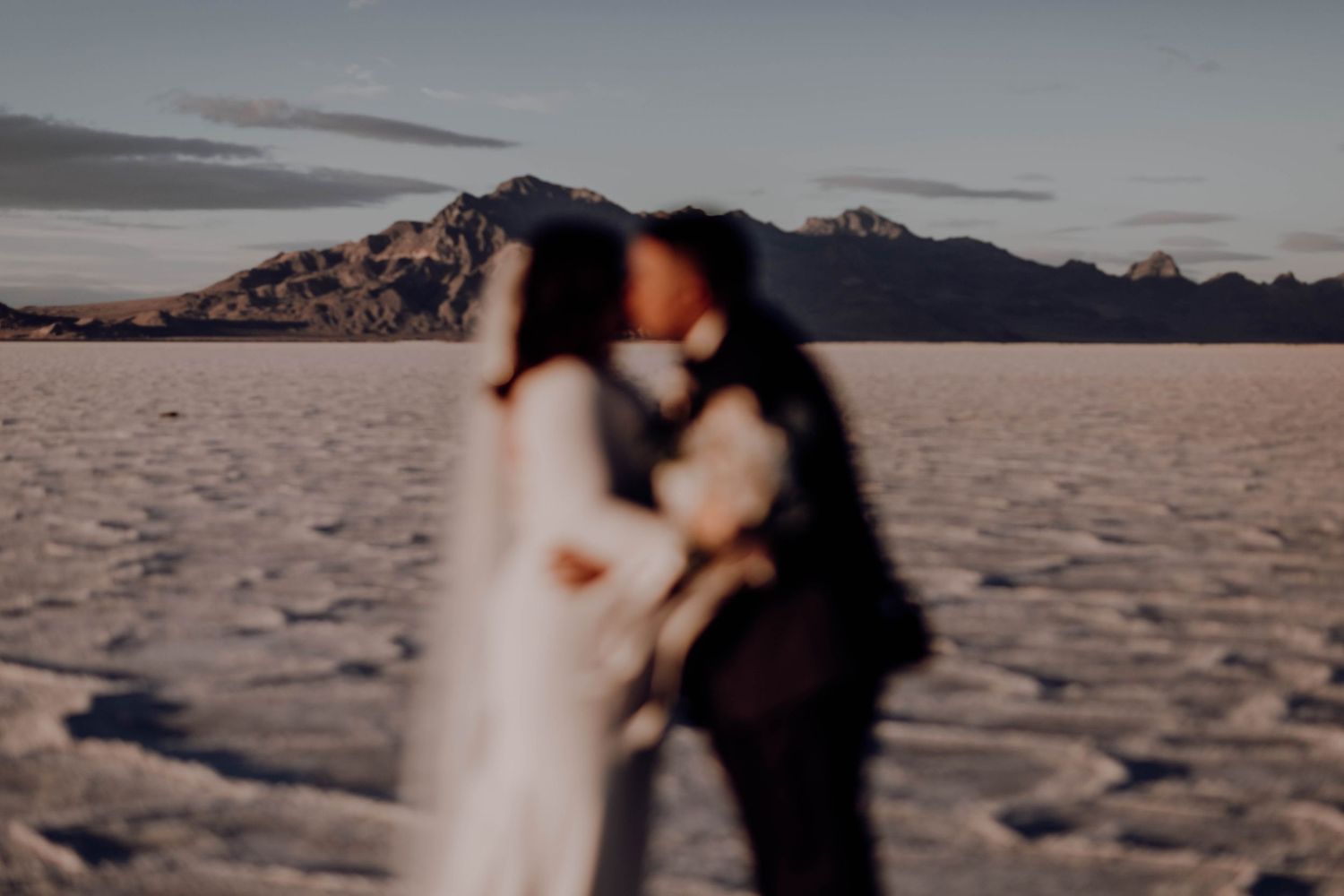 Couple sharing a romantic kiss on the vast Bonneville Salt Flats with mountain backdrop at dusk.