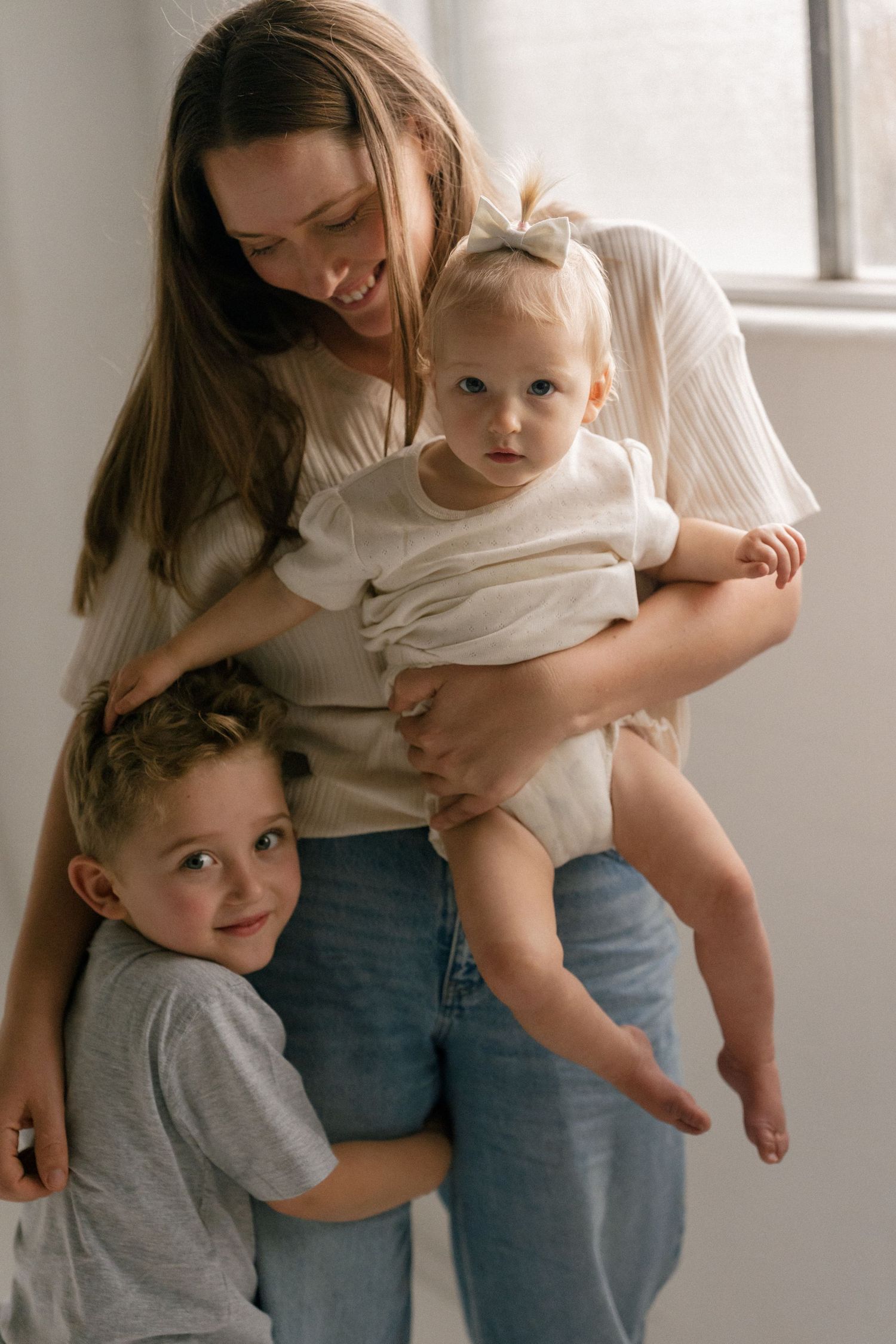 Smiling mother holds baby girl while young son hugs her legs near bright window.