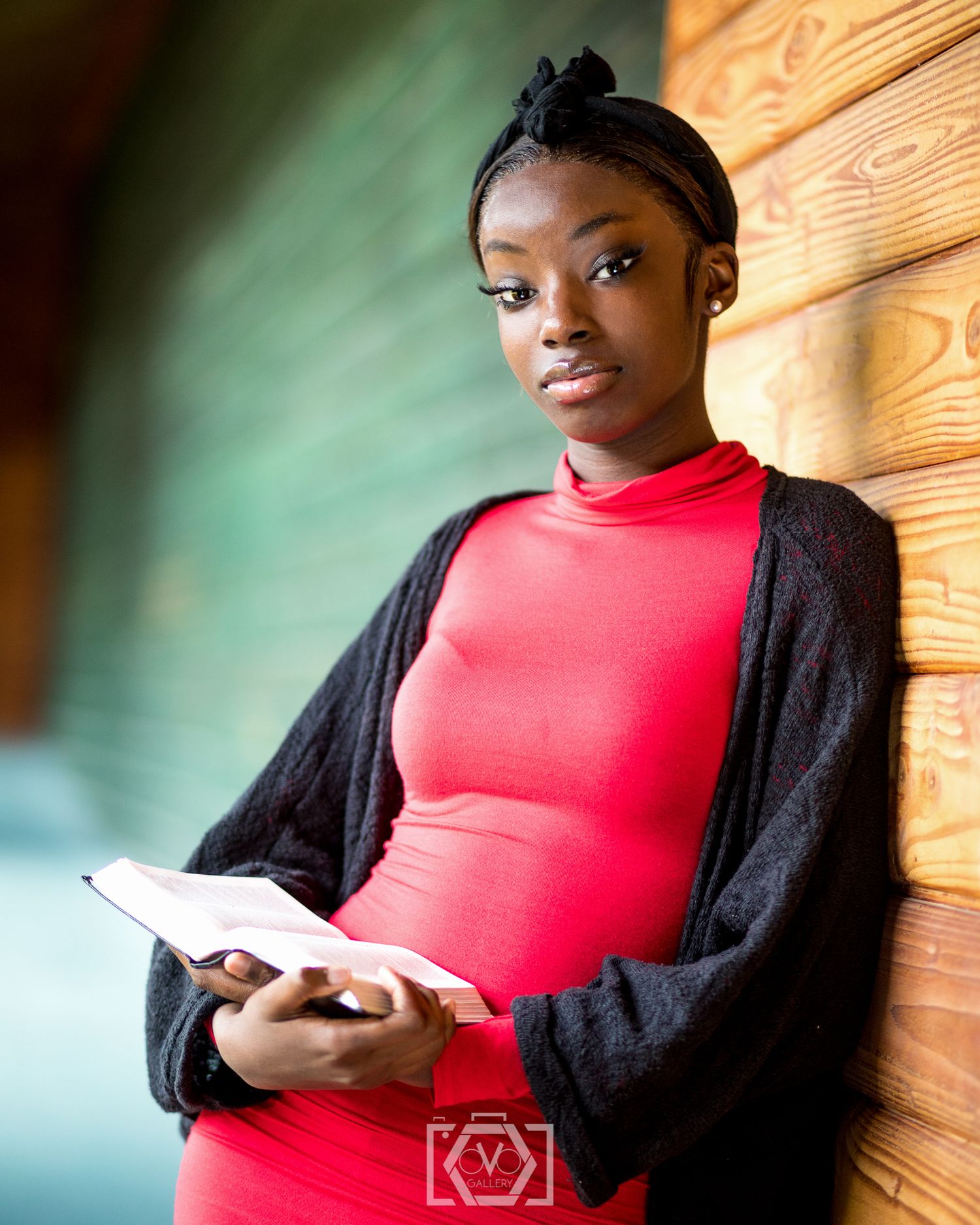 Young woman in red turtleneck dress and black cardigan holding a book while leaning against a wooden wall.