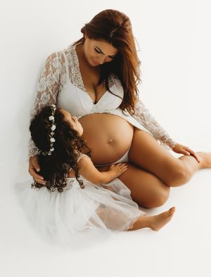A pregnant woman in white lace lingerie sits with a young child wearing a white dress on a light background.