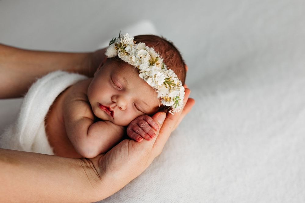 A sleeping newborn wearing a delicate white floral crown rests peacefully in gentle hands on a soft white blanket.