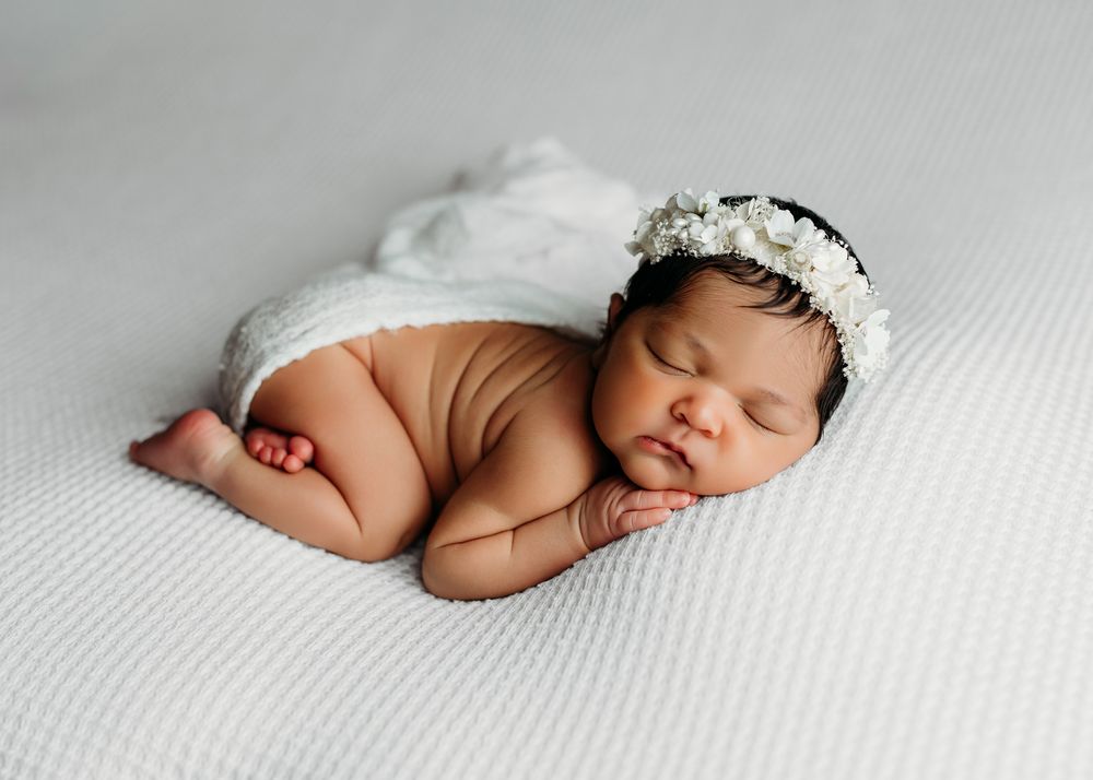 A sleeping newborn baby wearing a delicate white lace bonnet rests peacefully on textured white fabric.