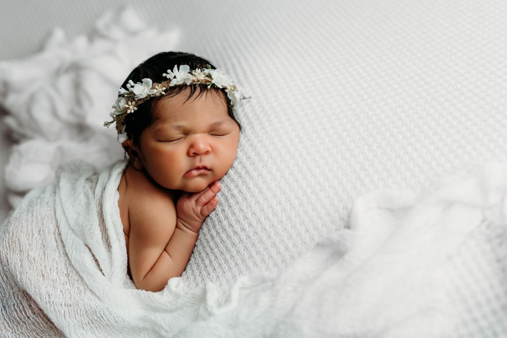 Newborn baby peacefully sleeping on white textured blanket wearing delicate floral crown.