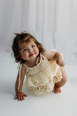 A toddler in a cream-colored lace dress crouches down on a white surface against a light gray background.