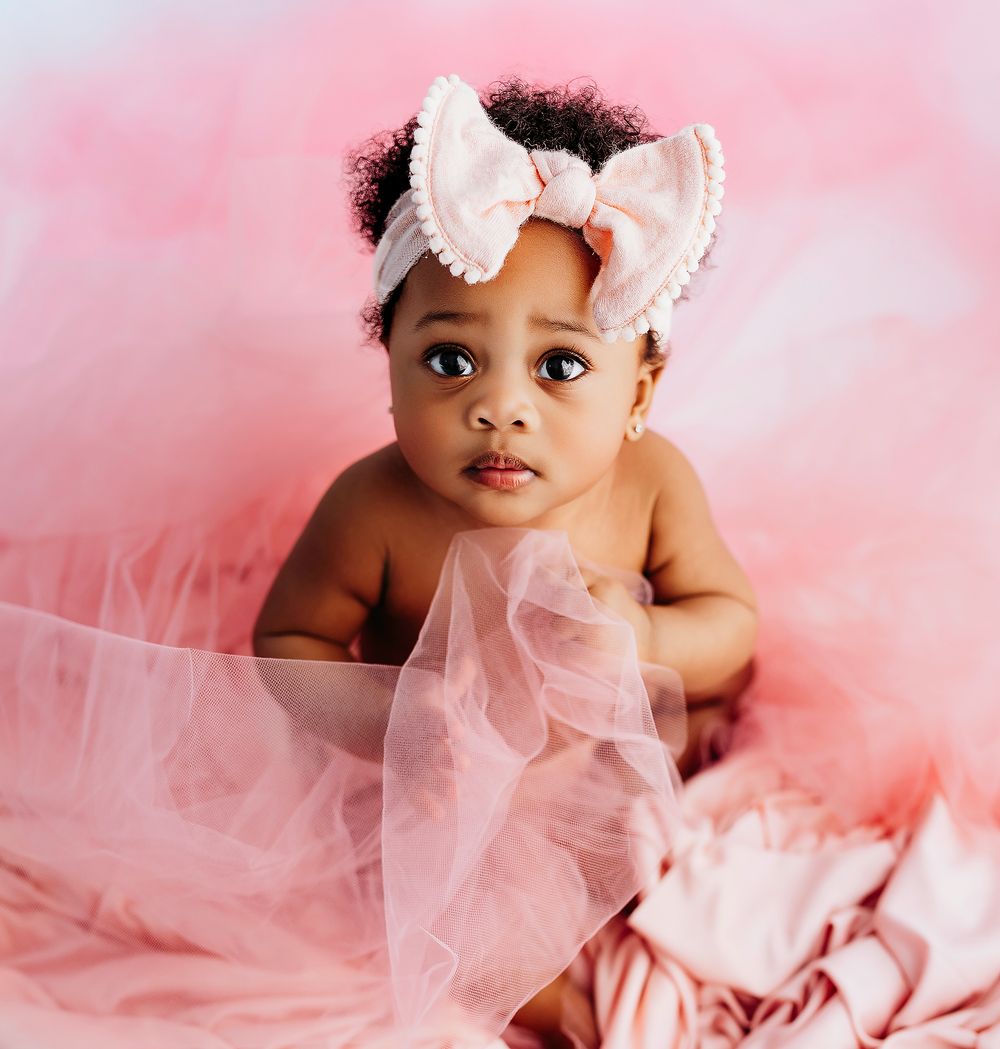 Baby wearing white bow headband sits amid pink tulle fabric against soft pink background.