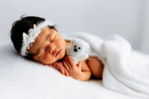 A sleeping newborn wearing a white lace headband and wrapped in a soft white blanket on light background.
Rashinphotography-Nova photographe