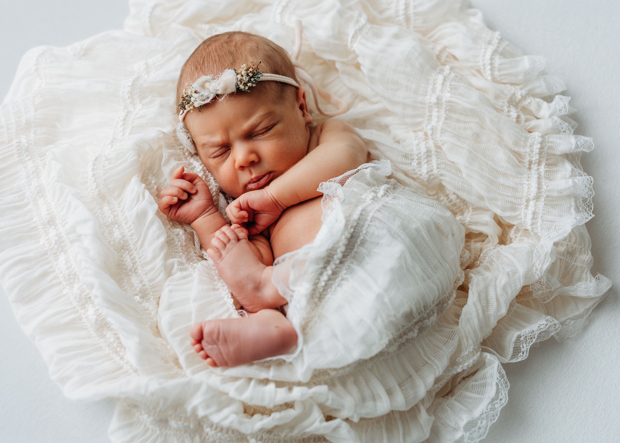 Newborn baby girl in white lace wrap and floral headband sleeping peacefully on ruffled cream blanket.