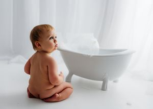 A baby sits beside a white bathtub in a bright setting with soft lighting and white decor.
