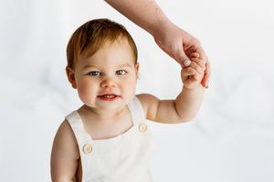 A small child wearing a white sleeveless outfit holds hands with an adult against a white background.