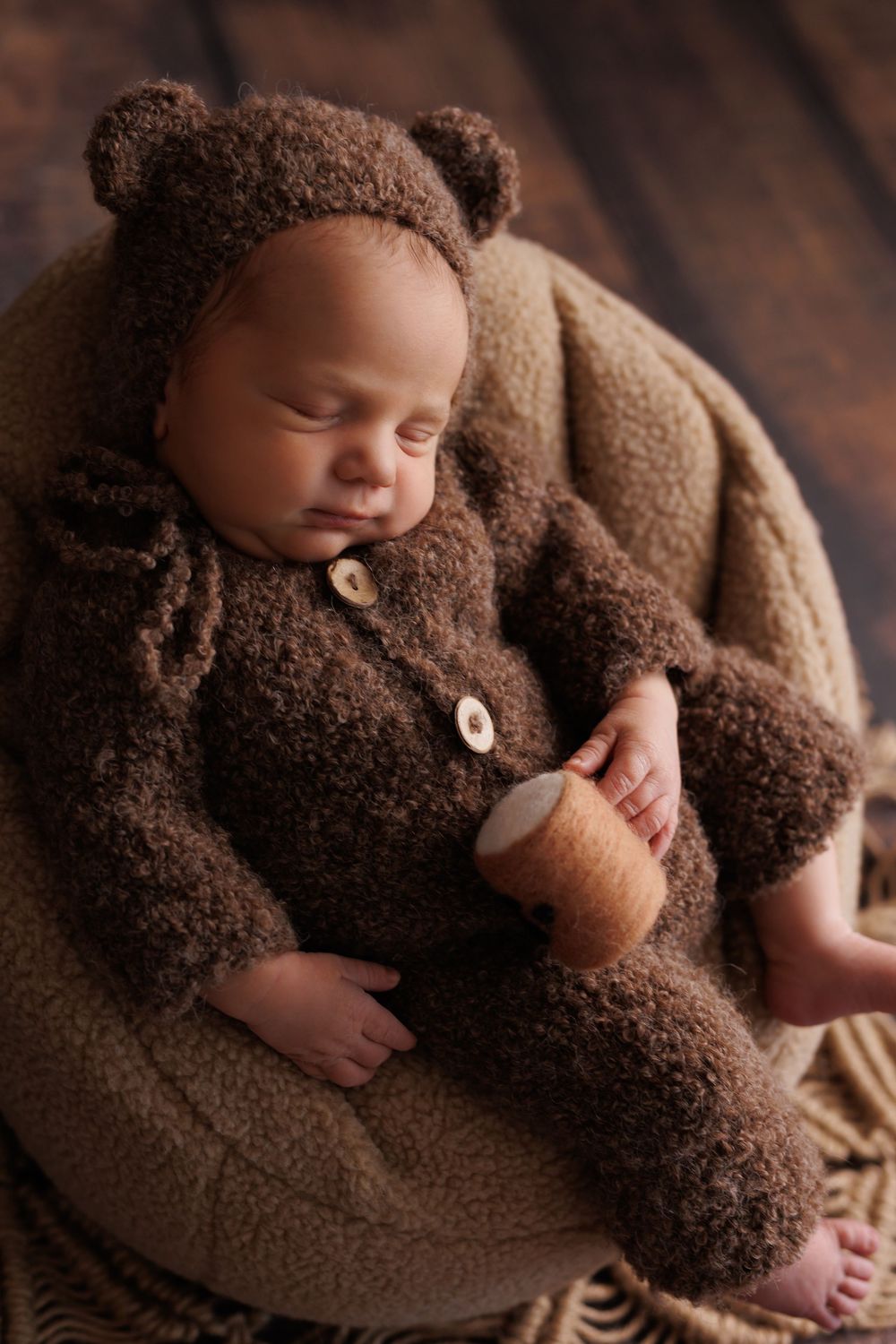 Sleeping baby wearing cozy brown teddy bear outfit curled up in beige blanket during newborn photoshoot.