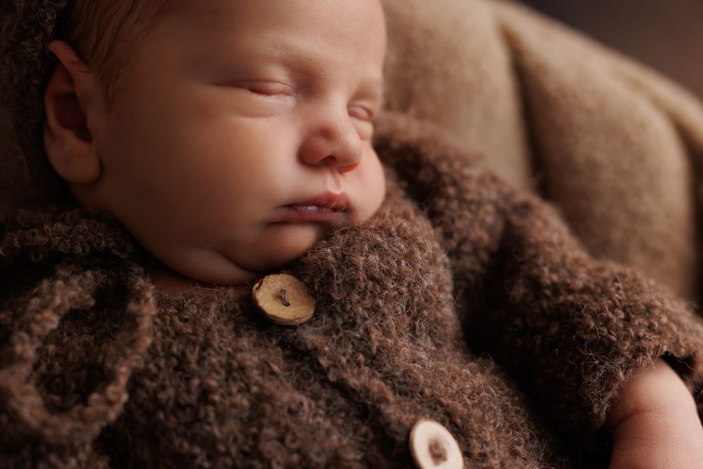 Sleeping newborn wrapped in brown textured blanket, photographed in soft lighting with a rustic wooden button accent.