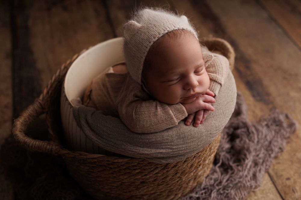 A sleeping newborn baby wrapped in soft brown fabric nestled in a cozy rustic wooden basket on a wooden floor.