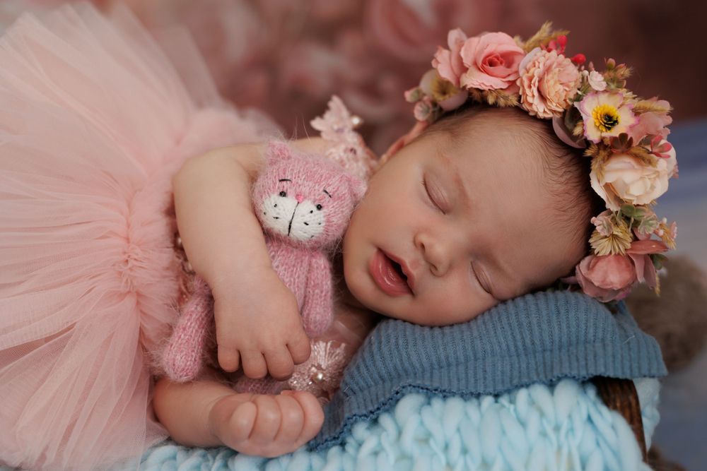 Sleeping infant in pink tulle dress and floral crown rests on blue blanket.