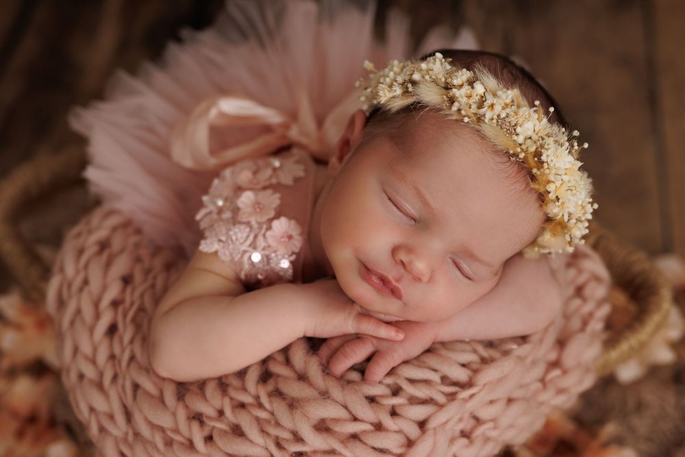 Newborn wearing flower crown sleeps on pink knitted blanket with delicate lace details.