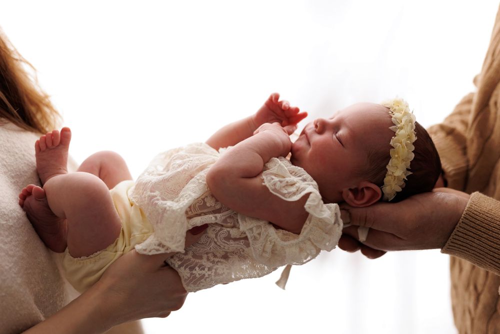 Newborn in white lace dress being gently cradled by caring hands in soft lighting.