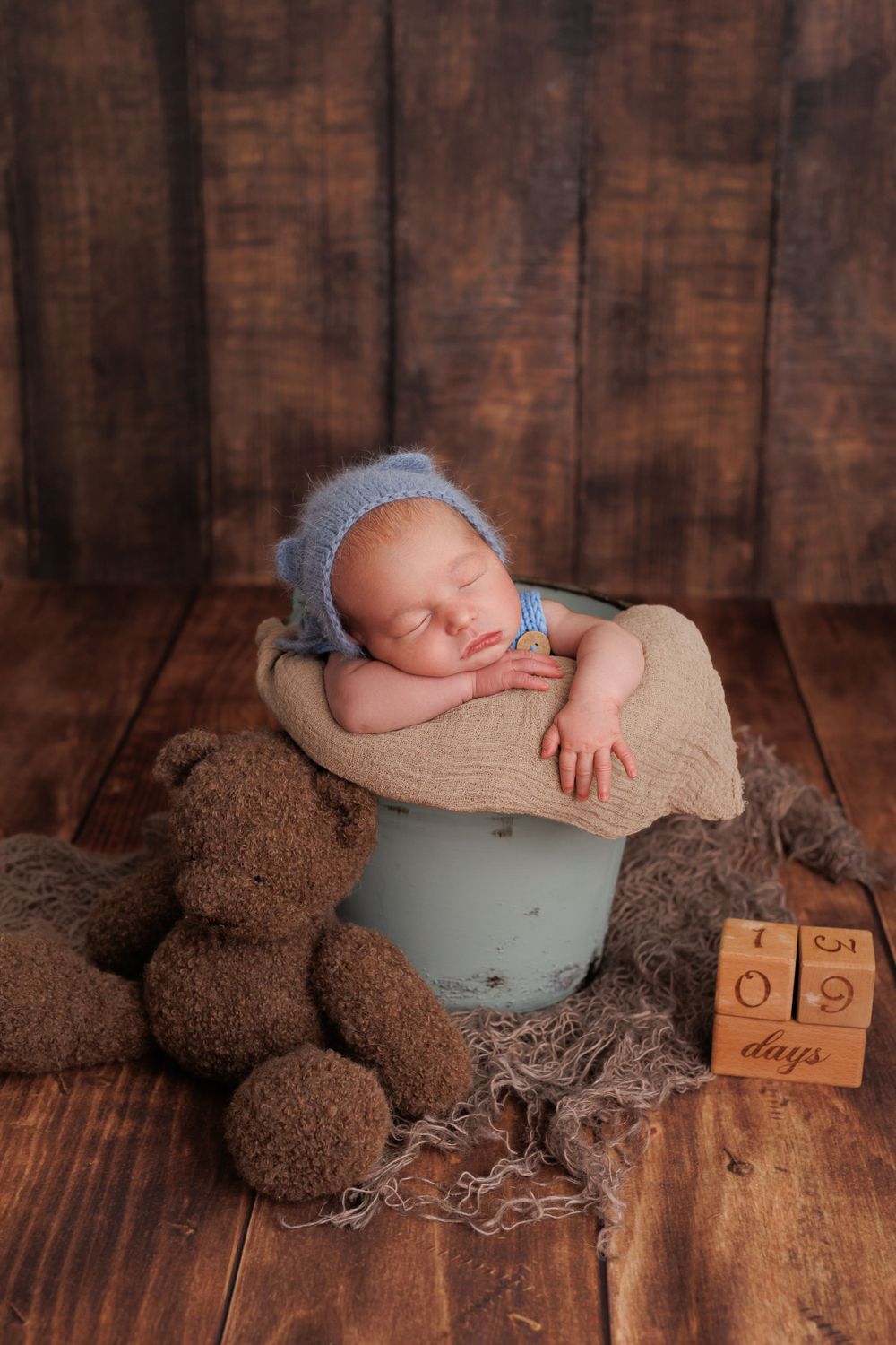 Sleeping newborn in grey bonnet rests on beige blanket with teddy bears and wooden blocks on rustic wood background.