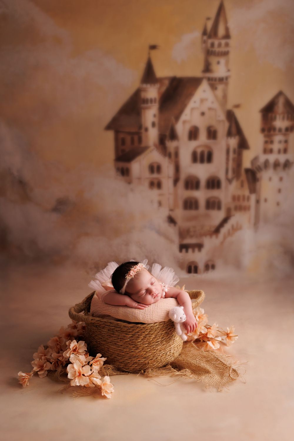 Newborn sleeping in woven basket against fairy tale castle backdrop with soft pink flowers.