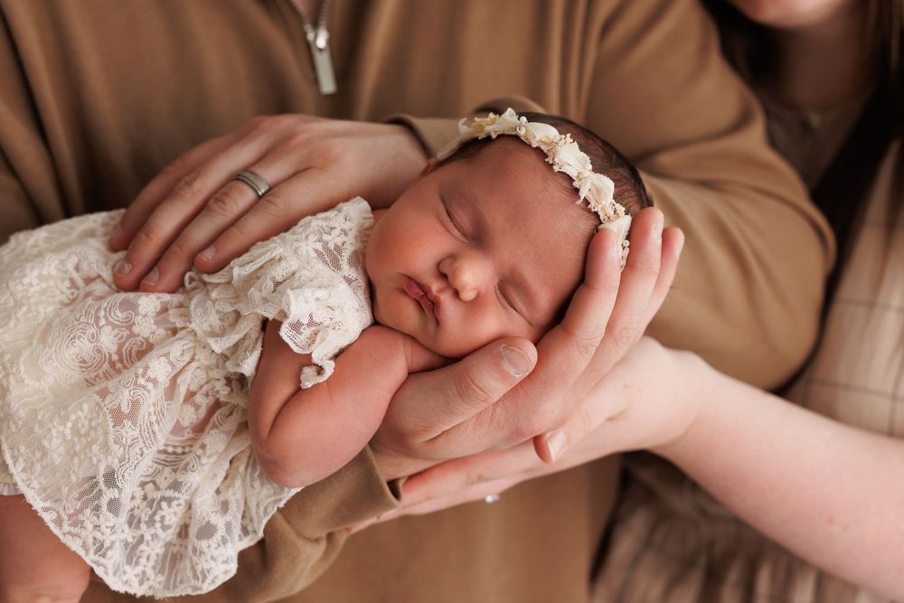 Sleeping newborn baby wearing white lace dress and floral headband rests in parent's hands.