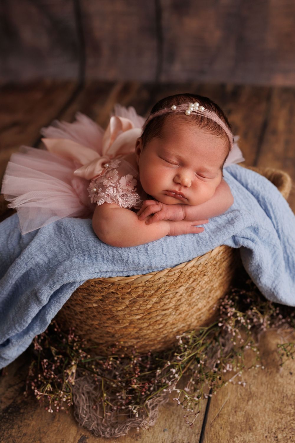 A sleeping baby in pink tulle rests peacefully in a rustic woven basket with light blue blanket on wooden surface.