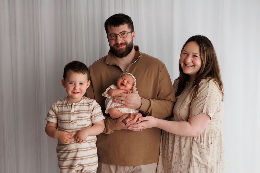 A family of four poses together against a gray backdrop in neutral-toned clothing for a portrait session.