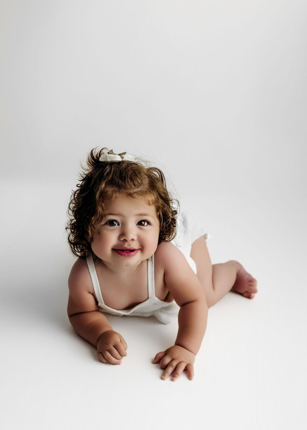 Baby wearing white top lies on stomach against light background, showing joyful expression with curly hair during a session in Fairfax, VA.