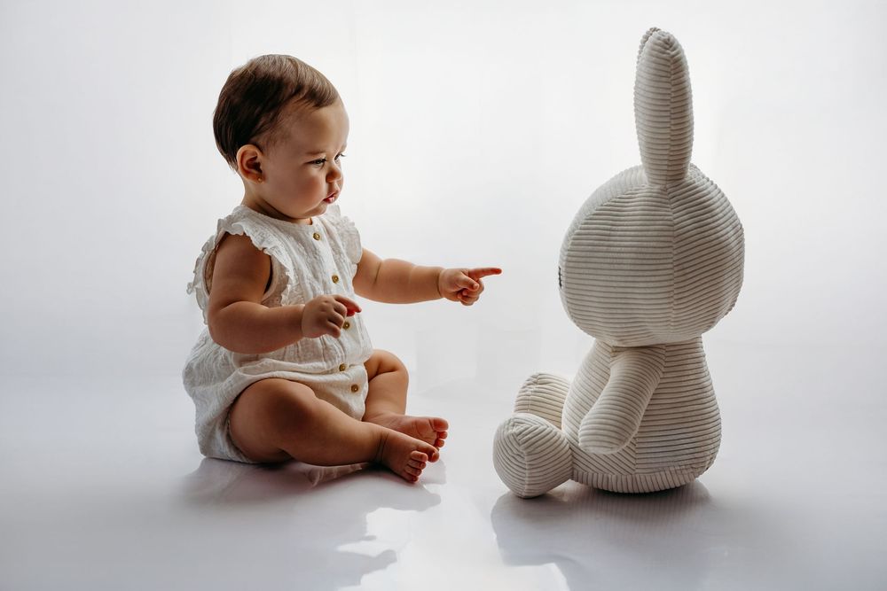 Baby in white outfit sitting beside large stuffed bunny toy on light background.
