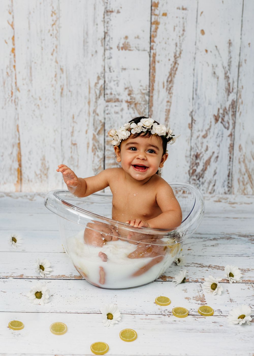 Smiling baby in a milk bath bowl wearing a floral crown, surrounded by daisies and gold coins on a white wood backdrop.