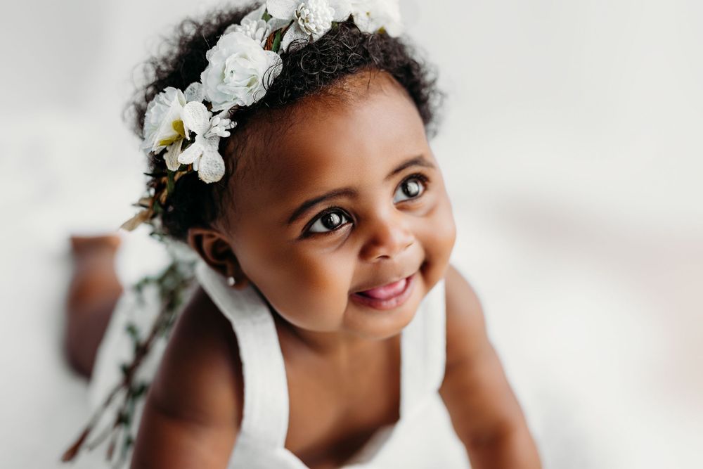 Sweet child wearing a floral crown smiles joyfully while wearing a white dress.