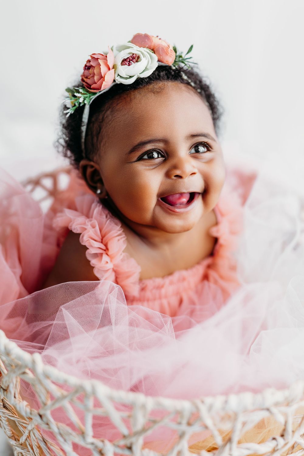 A baby wearing a pink tulle dress and flower crown smiles while laying on white blankets.