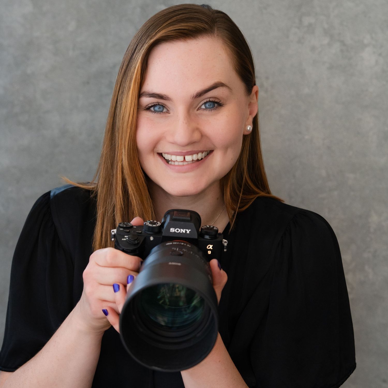 A photographer in a light top holding a professional camera against a dark background.