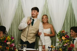 Groom smiles holding microphone while bride laughs holding his arm.