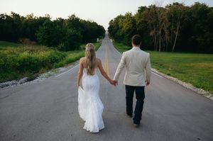 Bride and groom walk holding hands down tree lined country highway.