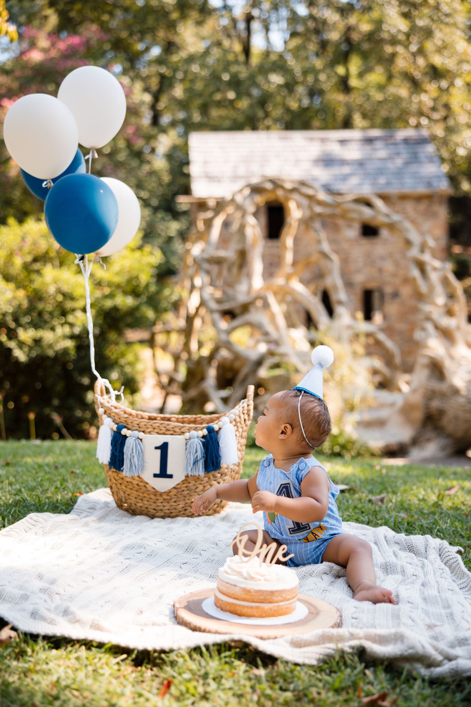 Outdoor cake smash photo session with blue and white balloons, white blanket, and wicker basket on grass.