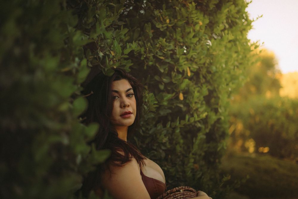 Portrait photography in golden sunlight shows figure with loose dark hair standing near lush green foliage at sunset in Redlands California