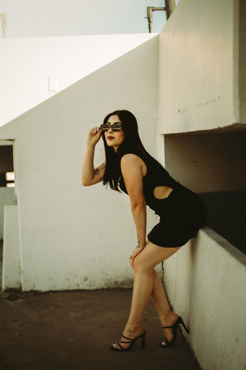 Model in a black dress and sunglasses poses against a white wall in an outdoor setting in Riverside California
