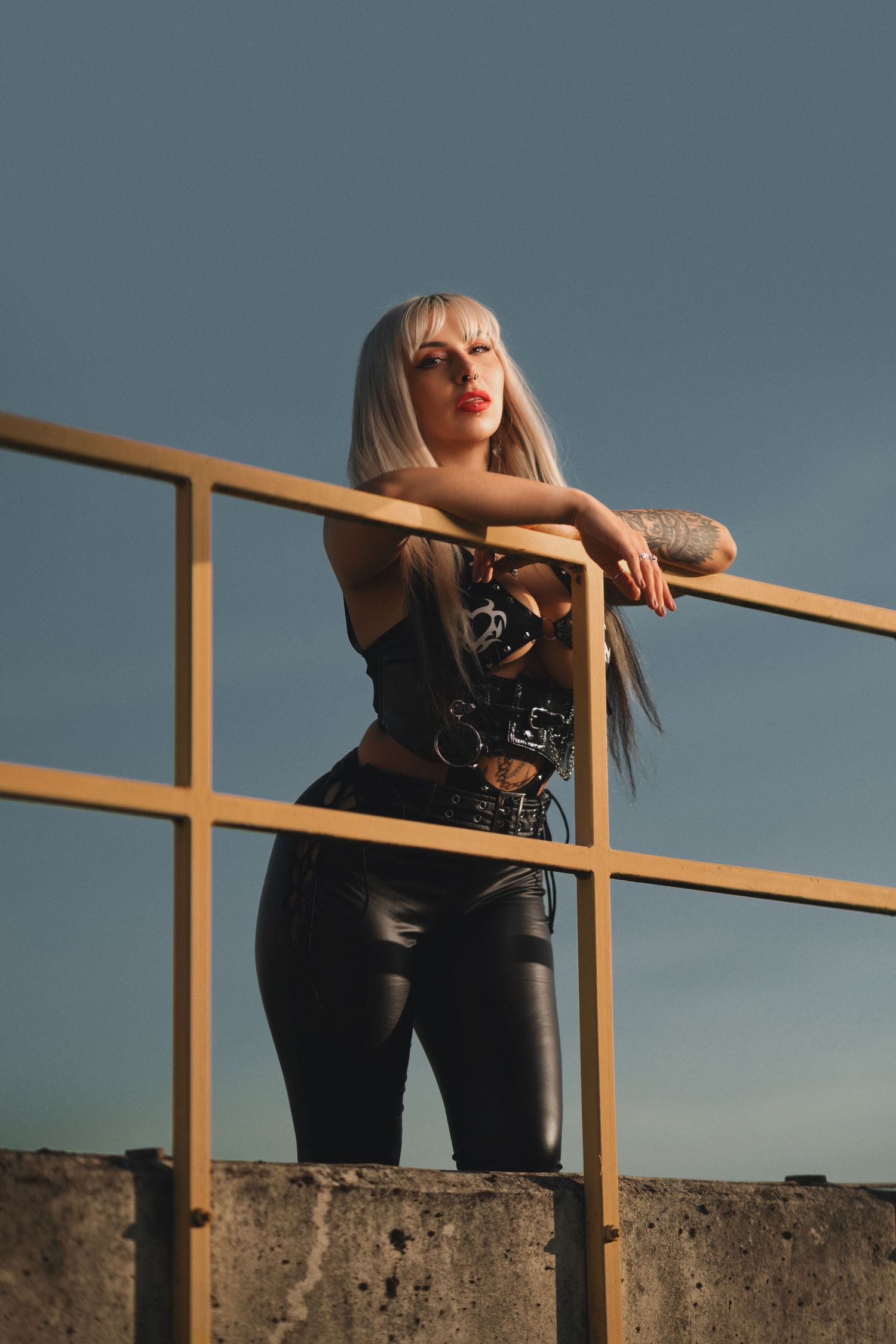 Tattooed blonde woman leaning on a metal railing in black leather, edgy alternative fashion portrait outdoors