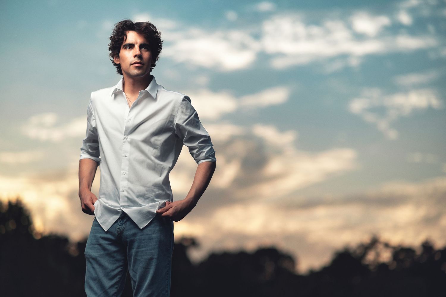 Young man with curly hair in a white shirt, three-quarter length environmental portrait at golden hour with dramatic sky