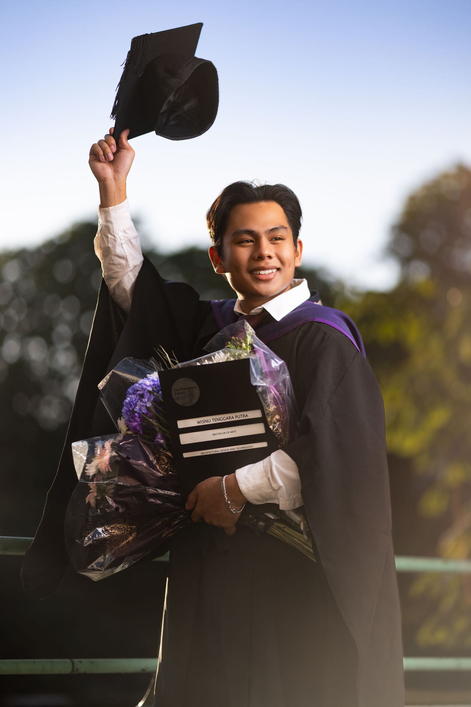 Graduate holding up mortarboard cap in purple robes with bouquet, outdoor graduation portrait photography