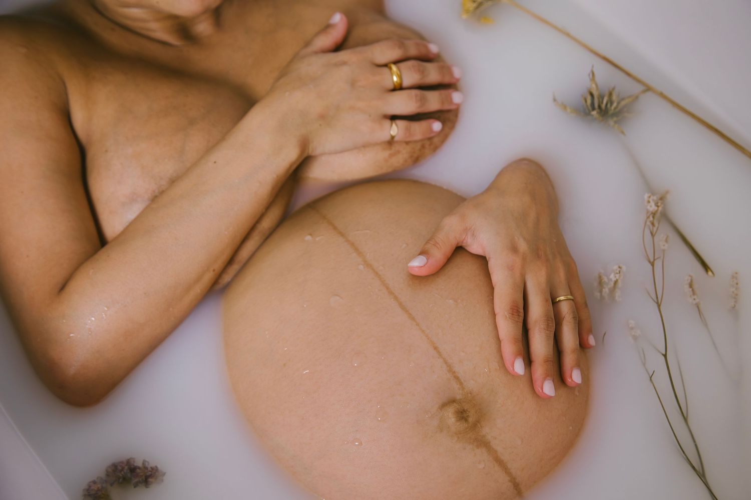 Gentle hands rest on a bare pregnant belly with dried flowers scattered around in a minimalist maternity photo.