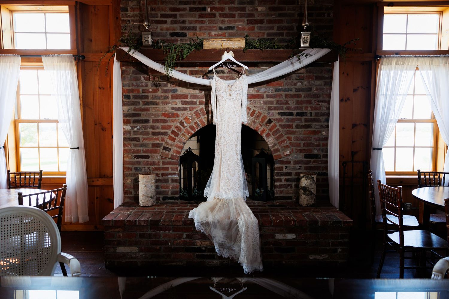 Elegant lace wedding gown displayed on hanger above rustic brick fireplace in barn venue.