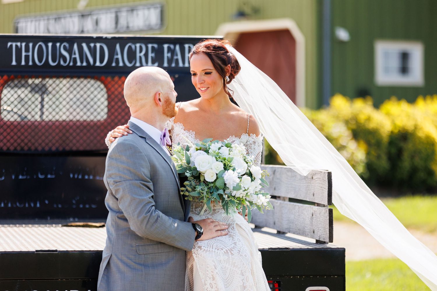 Bride and groom embrace near Thousand Acre Farm sign during outdoor wedding portrait session.