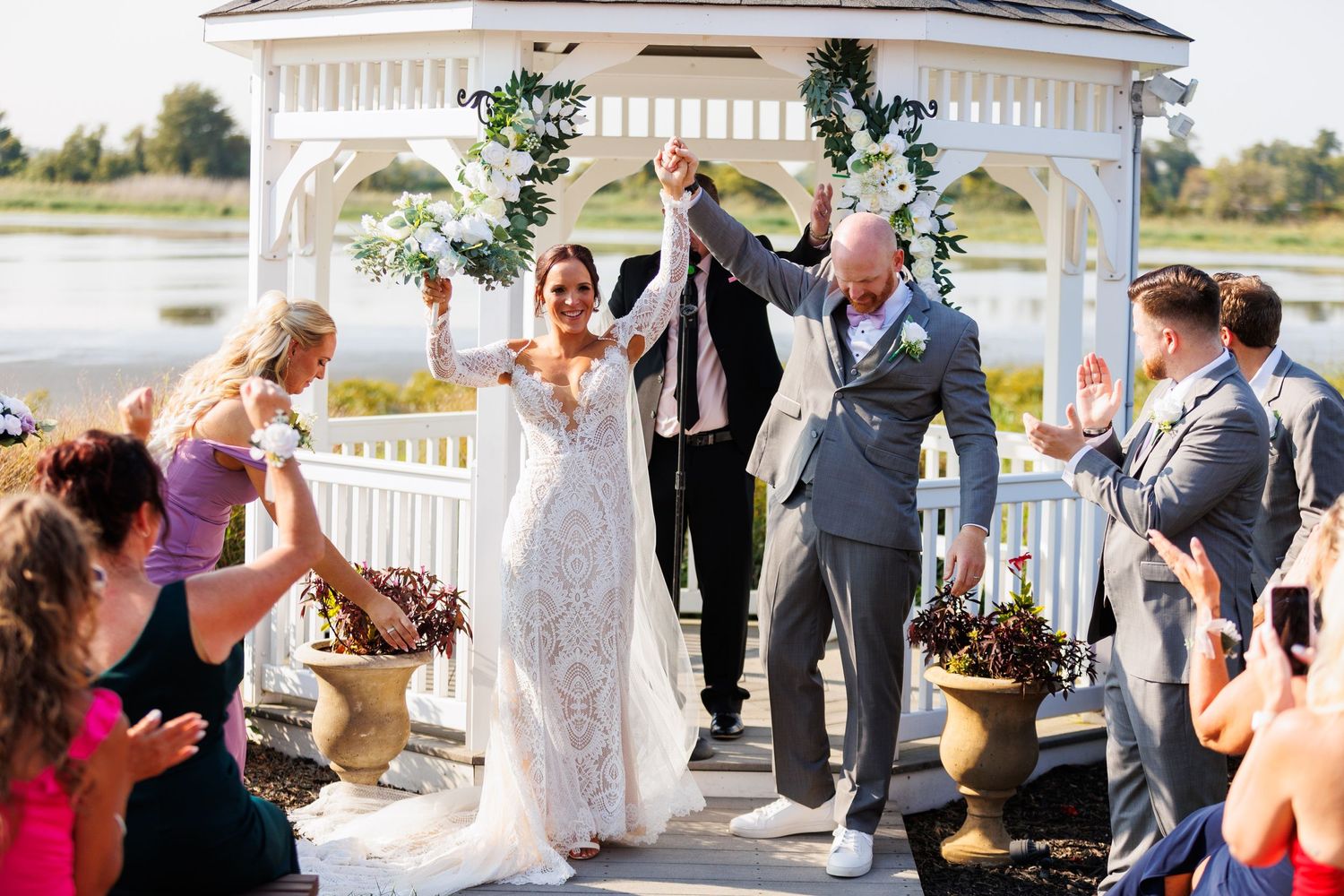 Newlyweds celebrate with raised hands exiting gazebo ceremony by waterfront as guests cheer.