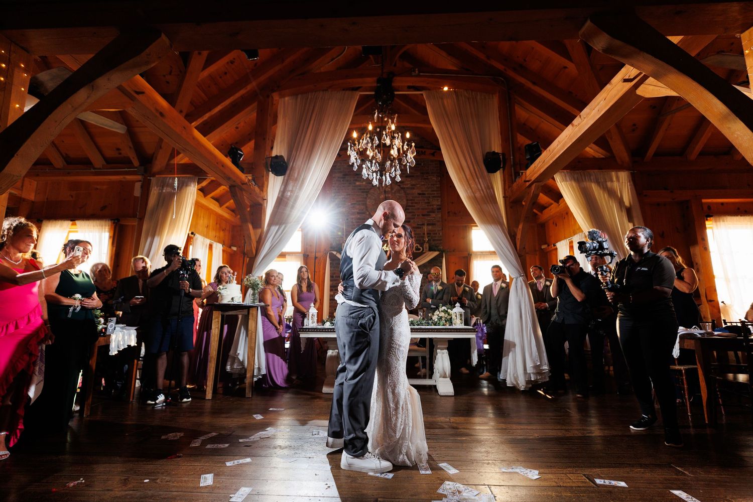 Bride and groom share romantic first dance under chandelier in stunning rustic barn wedding venue.