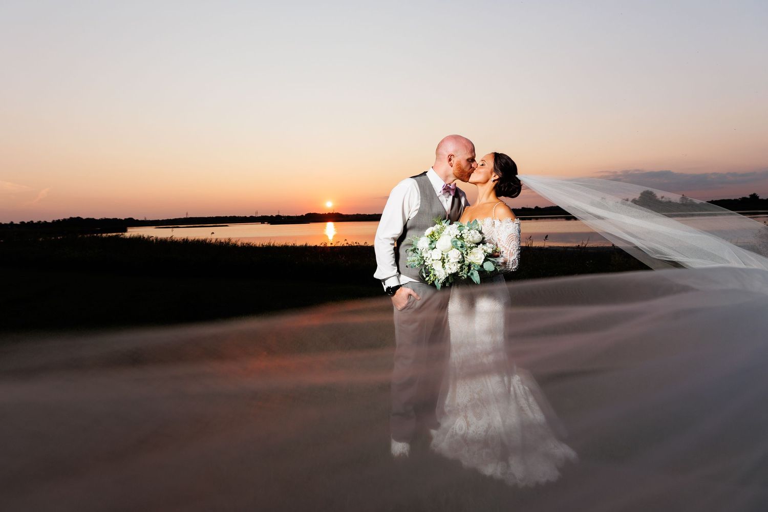 Bride and groom share sunset kiss on reflective beach with stunning orange sky backdrop.
