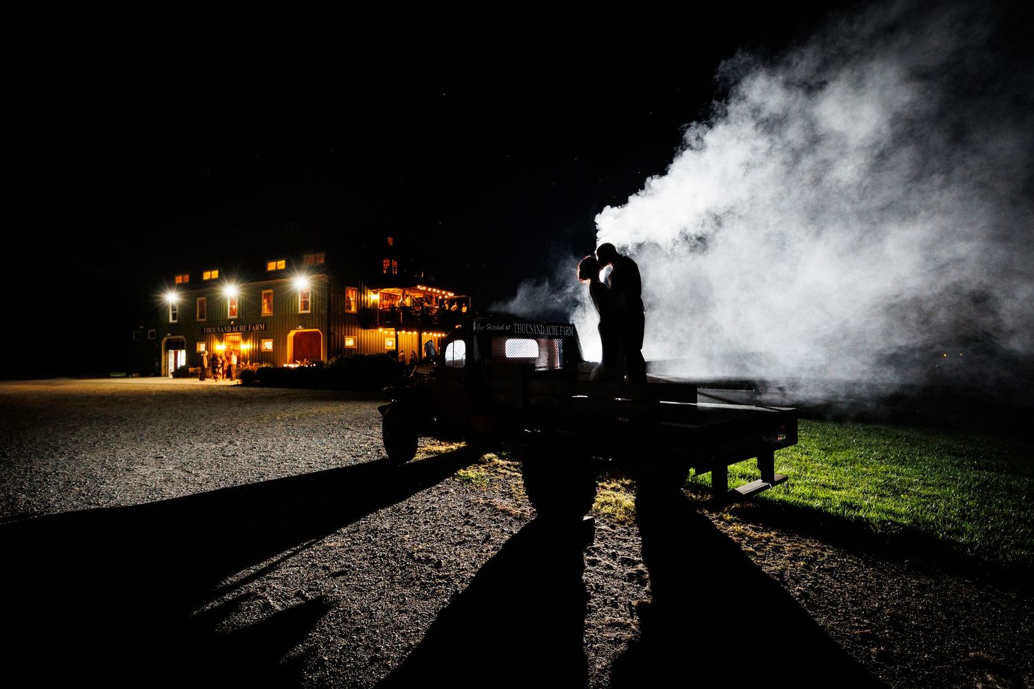 Silhouetted figure stands on truck bed with dramatic smoke against illuminated building at night.