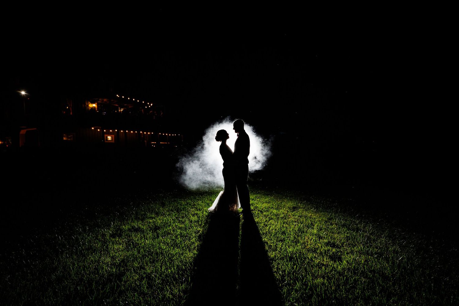 Romantic bride and groom silhouette surrounded by smoke on dark grassy field at night.