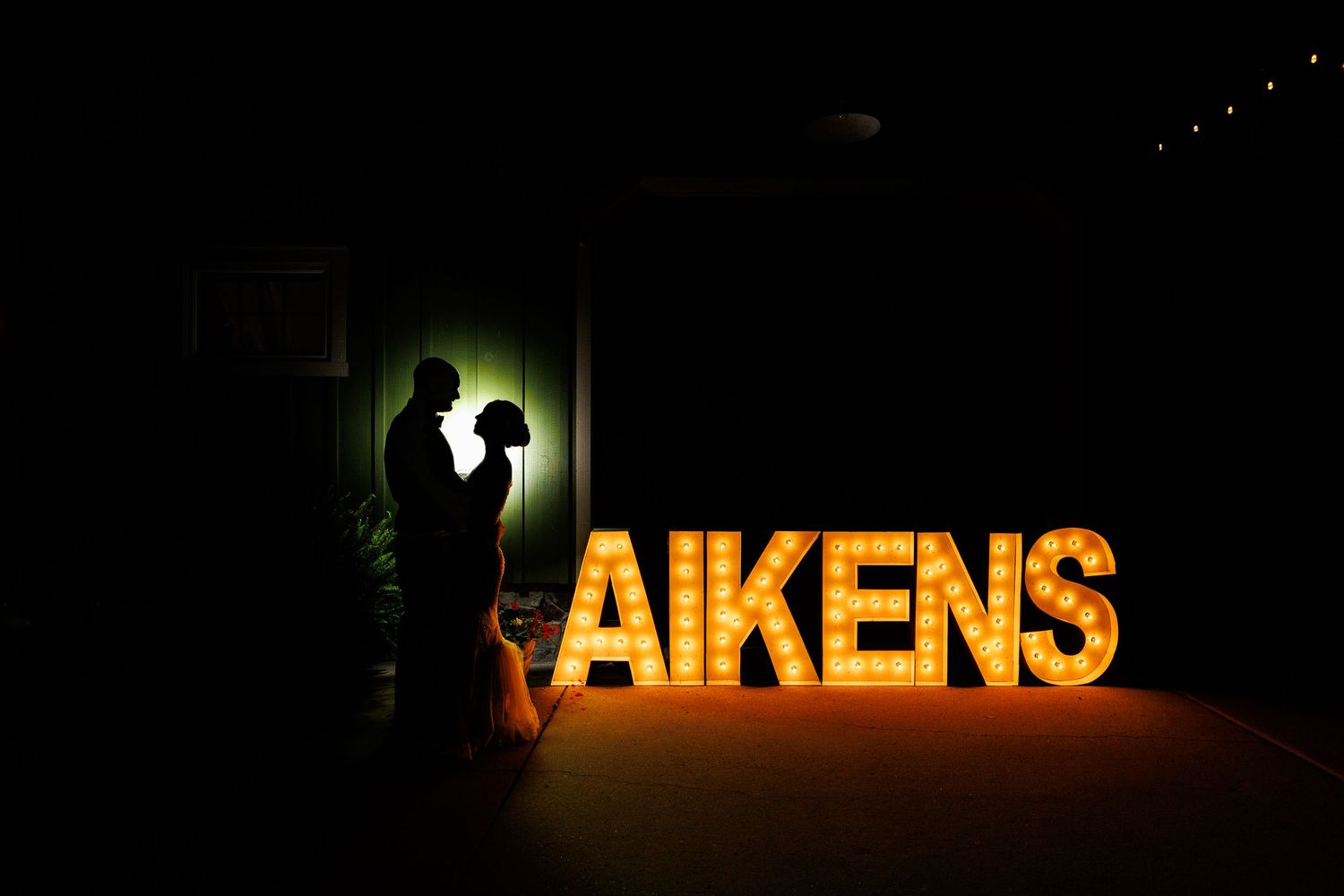 Bride and groom silhouette beside illuminated marquee AIKENS letters at outdoor night wedding.